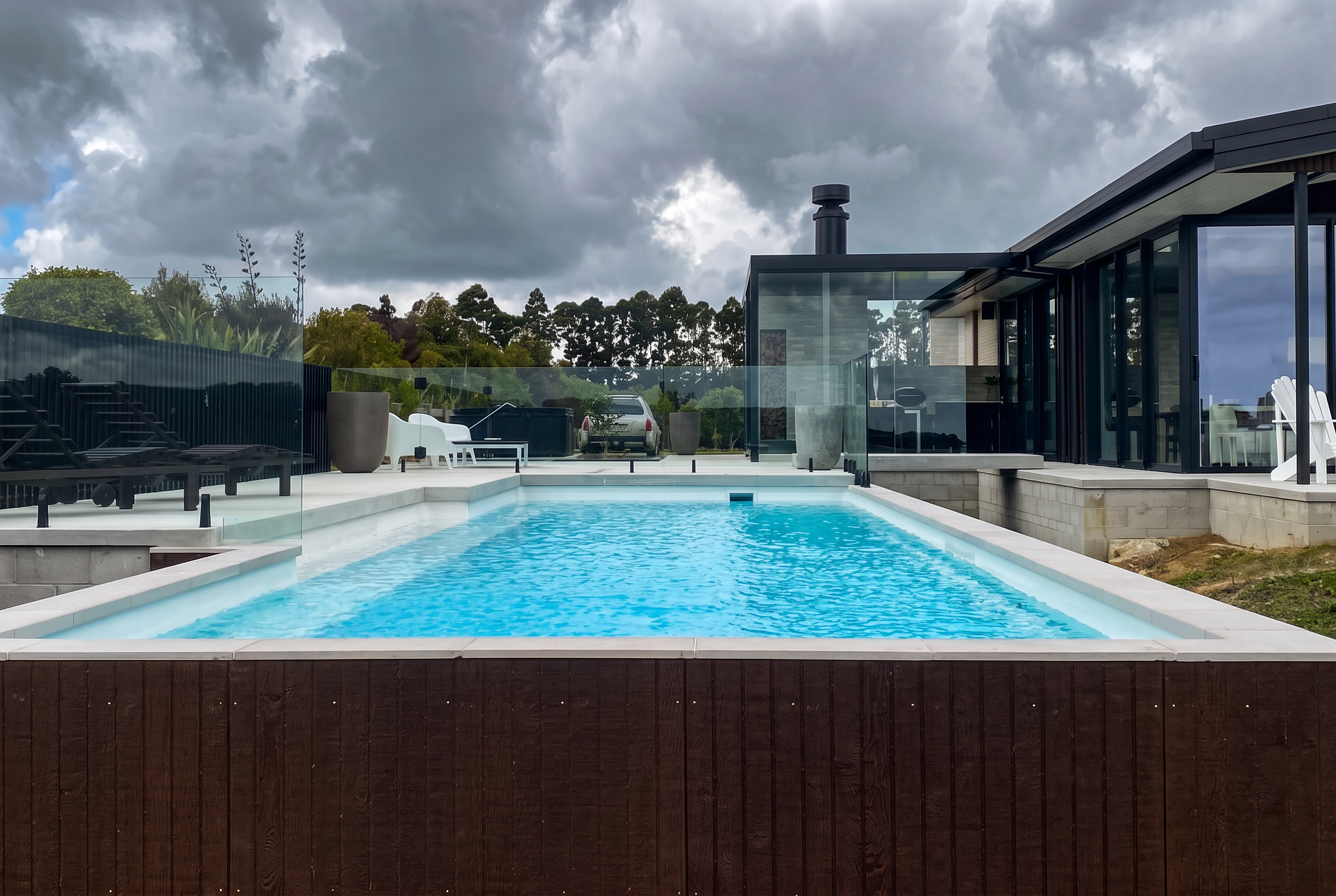 Pool with white coping and dark timber cladding beside a modern home